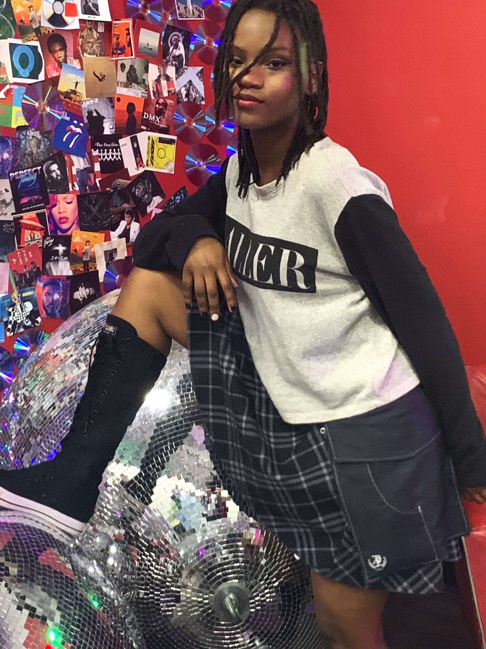 A young woman wearing a pair of midnight denim pockets with her foot on a disco ball, woth photos of music albums in the background